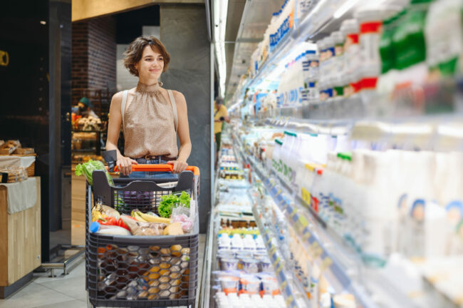 Young smiling happy caucasian woman 20s wear casual clothes shopping at supermaket store with grocery cart buy choose milk dairy produce inside hypermarket. People purchasing gastronomy food concept