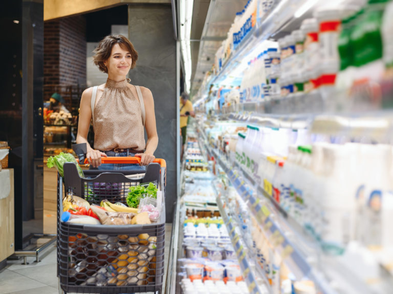 Young smiling happy caucasian woman 20s wear casual clothes shopping at supermaket store with grocery cart buy choose milk dairy produce inside hypermarket. People purchasing gastronomy food concept